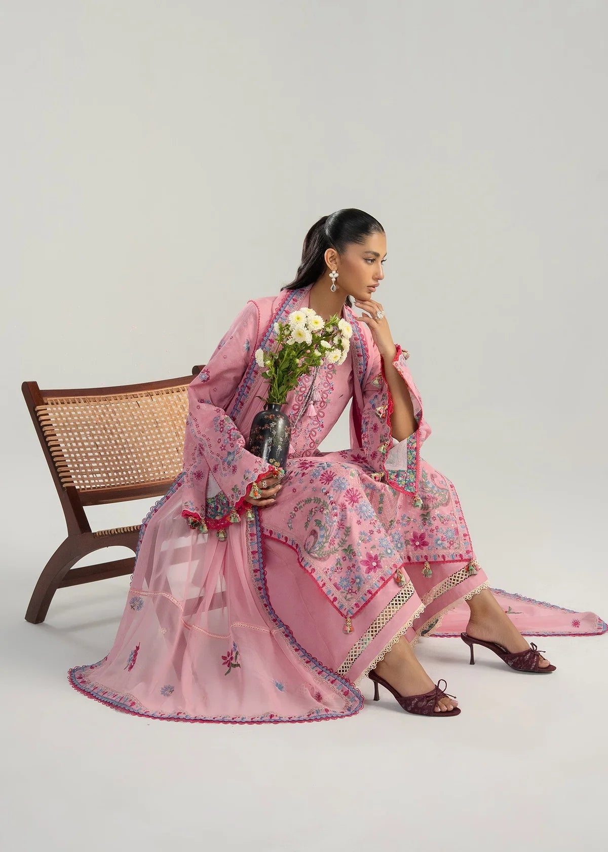 Sitting model presentation on a wicker bench highlighting the multi-panel mirror style pink and blue embroidery layout holding white daisies.